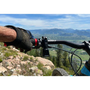 Bicycle handlebar with a gloved hand gripping the brake lever, set against a scenic mountain backdrop.