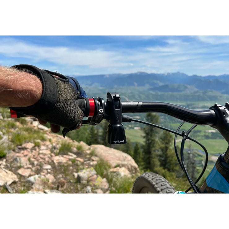 Bicycle handlebar with a gloved hand gripping the brake lever, set against a scenic mountain backdrop.